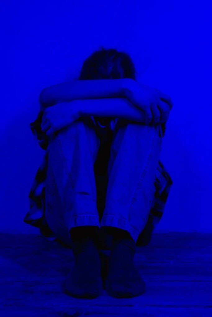 A person sits alone in a dark blue light, conveying feelings of sadness and solitude.